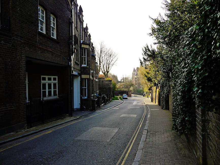 A narrow residential street with a paved, slightly curved roadway lined by a low brick wall and hedges on the right side, and a sidewalk with a metal fence and trees on the left. In the background, a 